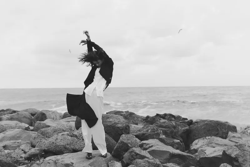 Rock, ocean, wind, movement, woman, dress, scarf, free spirit on a windy shoreline, black and white scene capturing motion and solitude as waves crash nearby