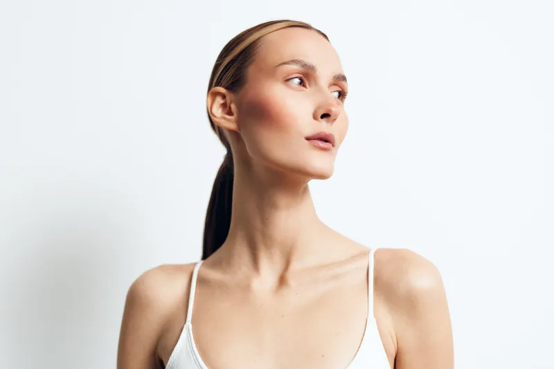 Young woman with clear skin and natural makeup looking to the side, wearing a white top, minimalist style, soft lighting, clean background, fresh and elegant appearance