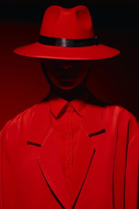 Model wearing bold red suit and matching wide brim hat stands in dramatic low light, creating mysterious fashion and beauty contrast effects characterized by intense shadows and color.