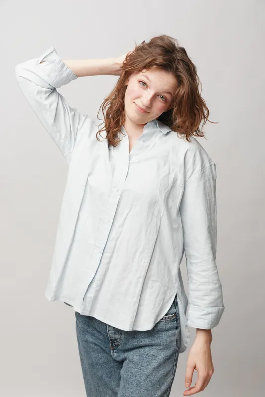 Young woman with curly hair in casual white shirt and jeans smiles gently, posing with hand behind head against plain light background. Concept of natural beauty and relaxed mood.