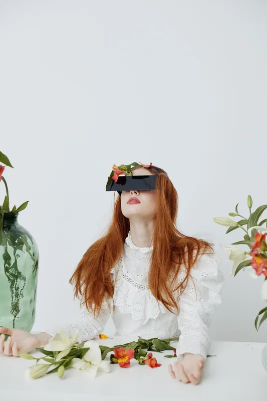 Artistic portrait of a model with long red hair wearing a vintage white dress, surrounded by flowers and greenery, conveying a serene and dreamy atmosphere