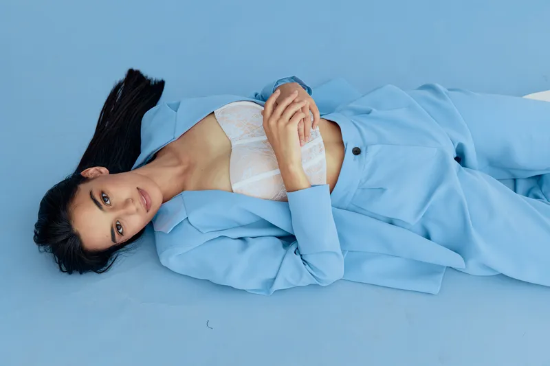 Young woman lying on blue floor wearing light blue suit and white lace top, posing with relaxed expression and crossed hands in minimal studio setting.