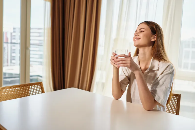Woman morning coffee at home by bright window, holding mug with smile and sunlight on face, relax with peaceful breakfast moment in cozy interior and soft natural light.