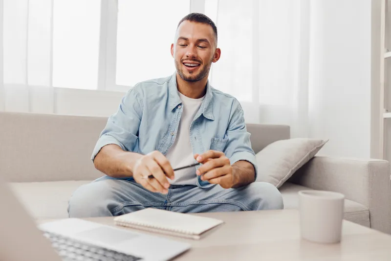 A smiling man engaging with his laptop at home, embodying concepts of emotional minimalism and work-life blur, with a cozy, light-filled atmosphere.