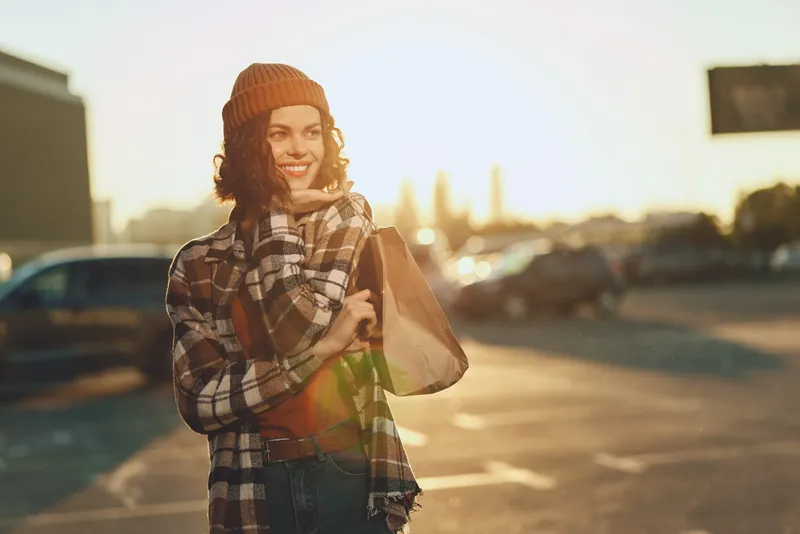 Woman shopping with smile at sunset in urban parking, candid portrait of beanie-wearing model holding shoppingbag. Authenticity, candid lifestyle, golden hour glow, mindful living.