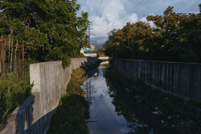 Urban canal with concrete channel and water reflection between trees under stormy sky, moody drainage scene with polluted banks and quiet late afternoon atmosphere for environmental stock.