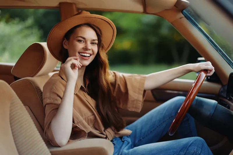 Smiling female model in stylish outfit, driving a car with a joyful expression, surrounded by green nature, representing freedom and adventure.