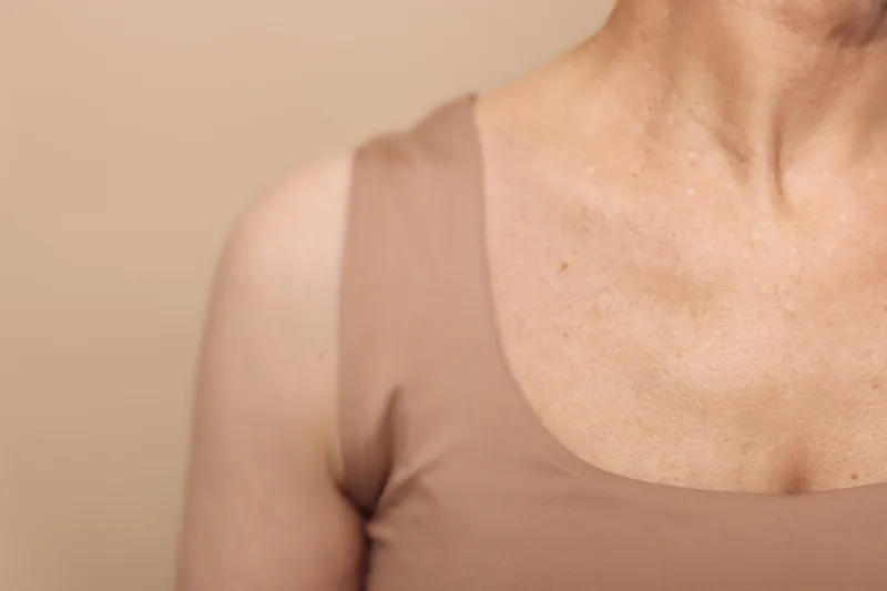 Close up of mature woman shoulder and neck with natural skin texture wearing beige sleeveless top against neutral background. Concept for aging, skin care, and natural beauty of older adults.