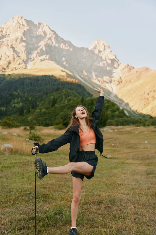 A joyful young woman stands in a rugged meadow, posing with one leg raised and a trekking pole beside her, under a dramatic alpine landscape with sunlit peaks.