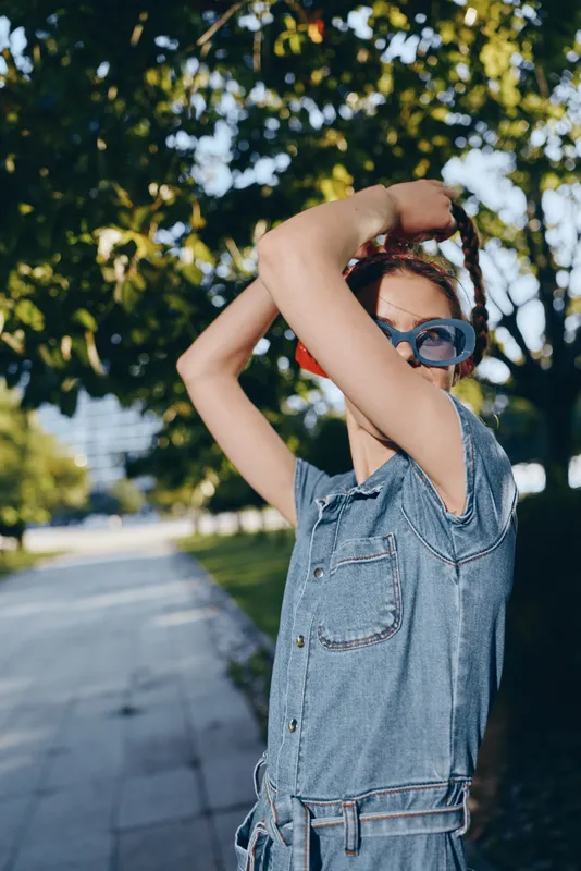 Young woman in denim jumpsuit adjusting sunglasses outdoors with bright sunlight and green trees in the background during warm summer day