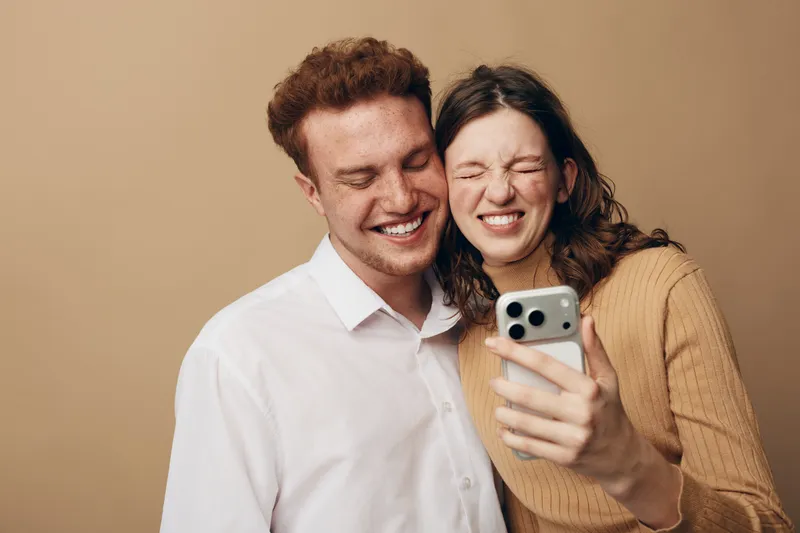 Happy young couple takes a selfie together with a smartphone. They smile and laugh in casual clothes against a beige background. Concept of friendship, technology, and joyful moments.