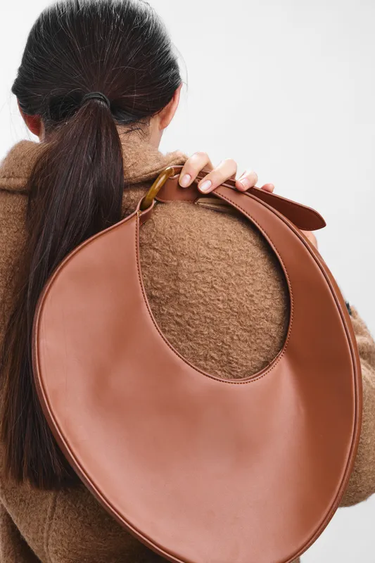 Asian woman stands with a circular brown leather bag slung over her shoulder, wearing a wool coat. The close view highlights texture, color, and contemporary urban style.
