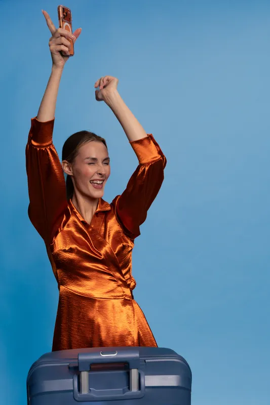 Happy young woman in orange dress celebrating with raised arms holding a phone behind a blue suitcase. Concept for travel, vacation, joy, and freedom on a light blue background.