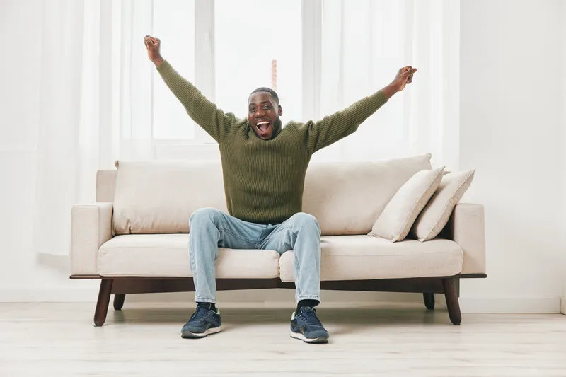 Happy African American man celebrating success while sitting on a couch in a bright, modern living room, wearing casual green sweater and jeans