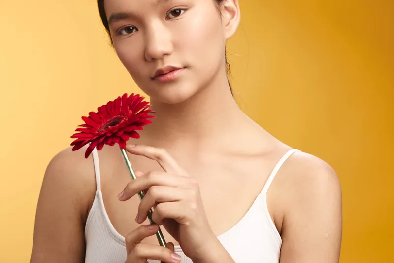 Asian woman stands against a warm yellow backdrop, gently holding a red flower. Her calm expression and soft pose convey elegance, poise, and a serene mood for lifestyle and beauty themes.