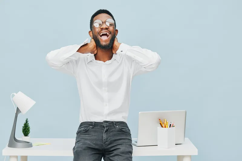 Joyful young man wearing glasses and a white shirt, standing behind a desk with a laptop, in a bright modern office space, expressing happiness and excitement. People lifestyle concept, positive emoti