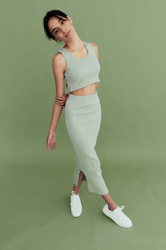 Young woman in stylish casual outfit posing gracefully against pastel green background, wearing crop top and midi skirt with minimalist white sneakers.