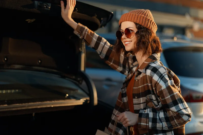 Woman at car trunk smile in plaid jacket and beanie with sunglasses, sunlight warming her face; candid lifestyle shot highlighting authenticity, golden hour glow, mindful living and emotional