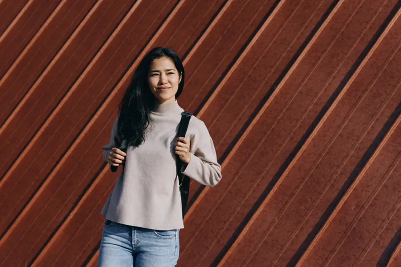 Casual city style with a backpack, a smiling woman stands before a diagonal wooden wall in a soft sweater and jeans, embodying urban fashion and travel readiness