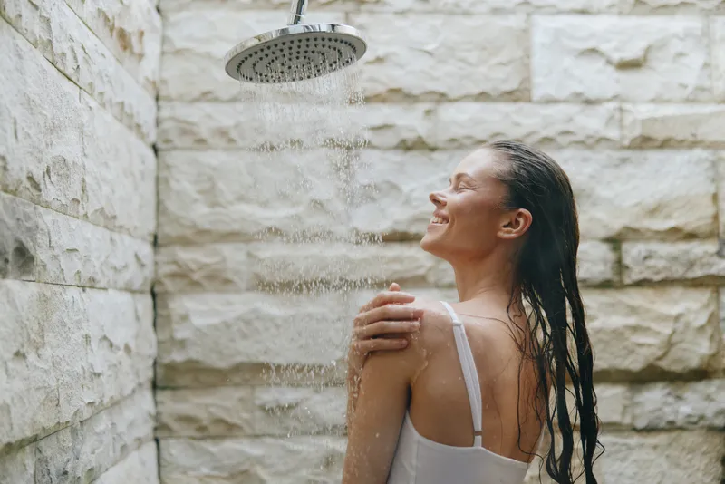 smiling woman enjoying outdoor shower with wet hair and relaxed expression near stone wall, natural light and refreshing water spray