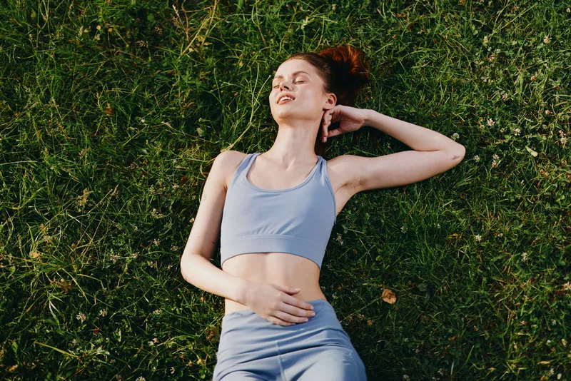 Young woman in athletic wear lies on green grass, enjoying a moment of relaxation and mindfulness, embodying wellness and healthy lifestyle concepts