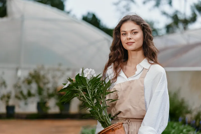 Young woman holding a potted flower, dressed in a beige apron and white shirt, smiling in a garden setting with greenhouses in the background, conveying a sense of peace and connection to nature