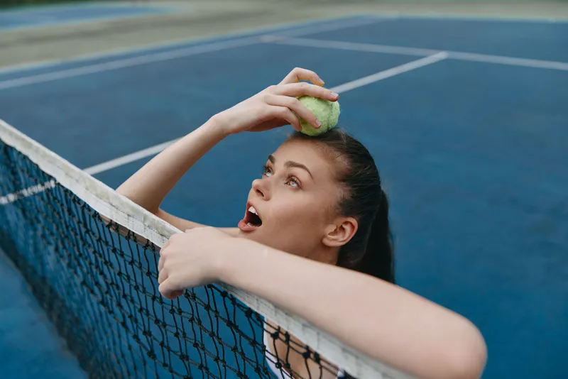 Young athletic woman surprised while playing tennis, holding a tennis ball above her head, energetic emotion captured on a blue tennis court background