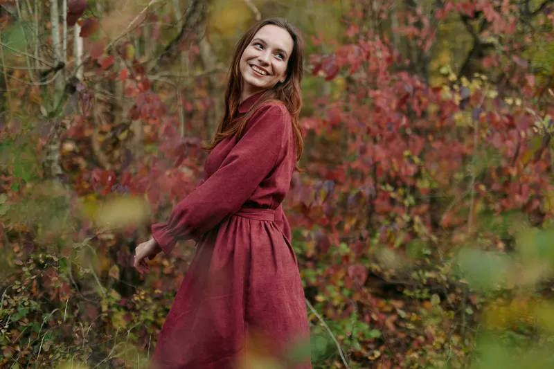 A young woman in a red dress smiles candidly among autumn foliage in a natural unposed portrait, conveying sincere connection, relaxed movement and true emotional presence.