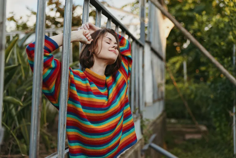 Woman in rainbow sweater leaning on metal railing, smiling with closed eyes. Young adult model in casual outfit with short brown hair, relaxed outdoor garden lifestyle portrait.