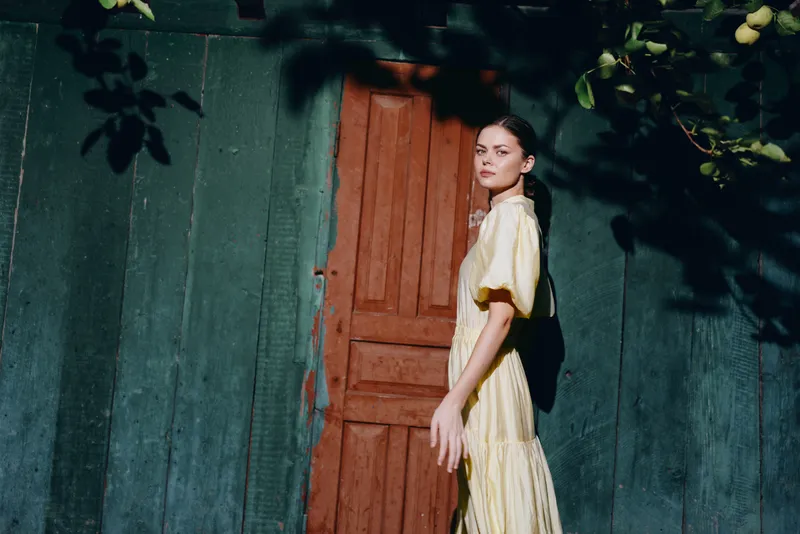 Portrait of a woman in a flowing yellow dress standing by a wooden doorway in a green wall setting, exuding calm elegance and summer mood.