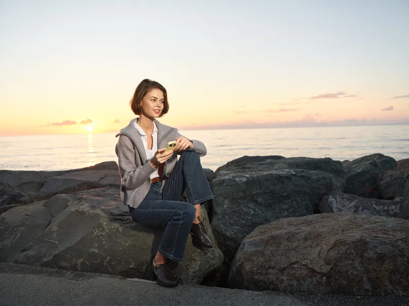 A professional woman sits on rocks by the sea at sunset, texting on her smartphone with a smile, dressed in blazer and jeans, projecting a calm, productive mood.