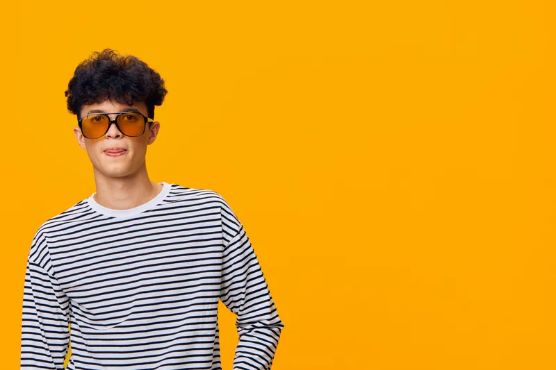 Young man wearing striped shirt and sunglasses posing confidently against vibrant orange background, showcasing relaxed style and modern fashion trends.