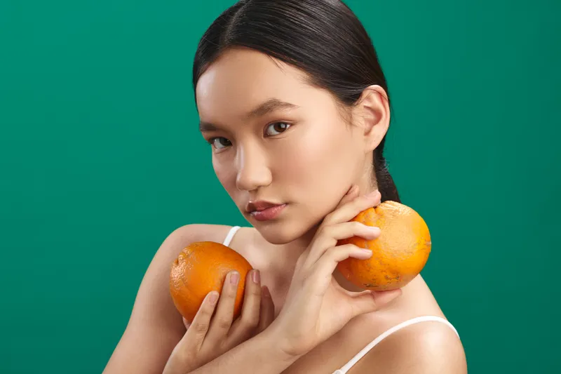 asian woman holds two oranges against a vibrant green backdrop, studio portrait with bold color and calm expression, creating a fresh still life that feels modern and artistic.