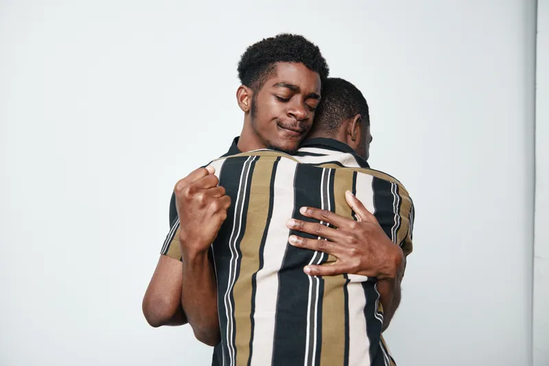 Two young black men hugging each other with eyes closed and peaceful expressions. Dark skin, casual clothing, emotional connection, friendship, support, and comfort in studio setting.