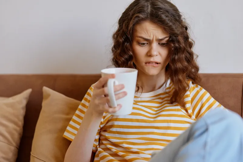 Woman with curly hair sits on a sofa holding a mug, looking thoughtful. Home living scene with striped shirt, cozy cushions, warm light, conveying lifestyle and comfort.