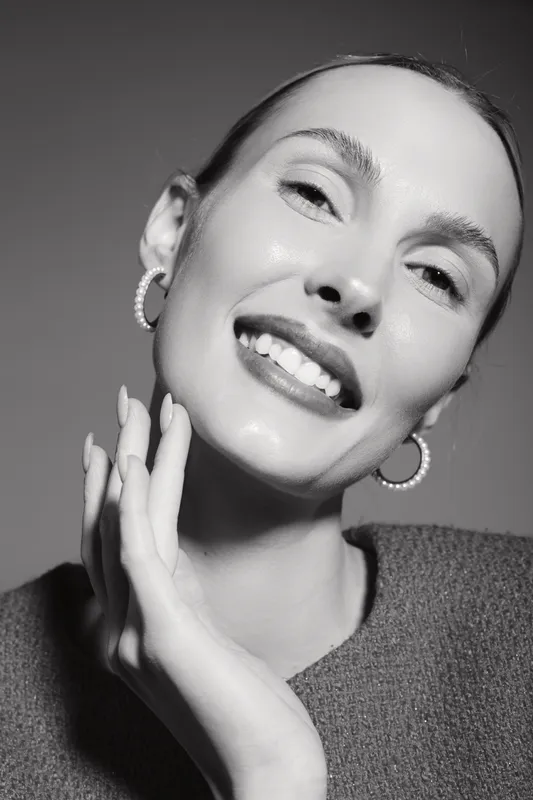 Smiling Caucasian woman with fair skin touching her face, wearing hoop earrings and knitted sweater. Concept of natural beauty and skincare.