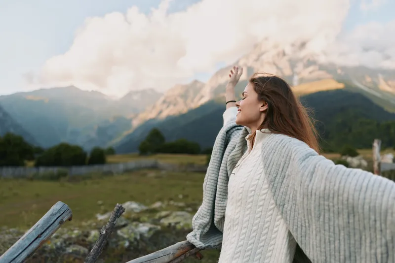 Young woman with long hair enjoys fresh mountain air while wrapped in a cozy gray shawl standing near a wooden fence with scenic peaks in the background.