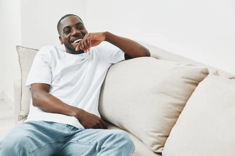 Smiling African American man in casual outfit sitting on a sofa, exuding happiness and relaxation in a light colored modern interior