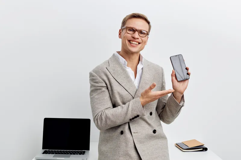 Smiling man in smart casual attire holding smartphone and pointing at it, with laptop and notebook on desk behind him, in bright modern office, casual business environment, technology, communication, 