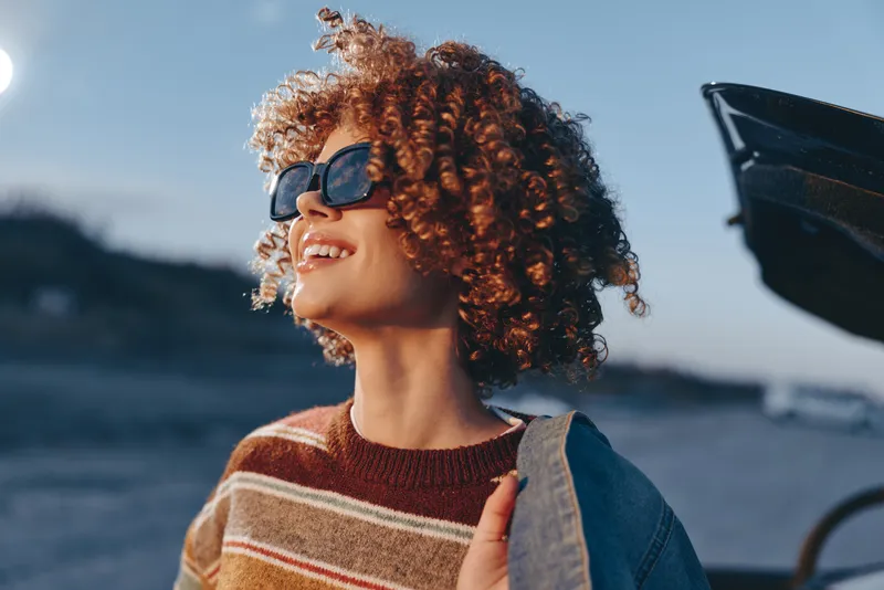 Woman lifestyle portrait, curly hair woman wearing sunglasses, smiling outdoors at the beach, holding denim jacket, striped sweater, summer sunlight, vacation mood