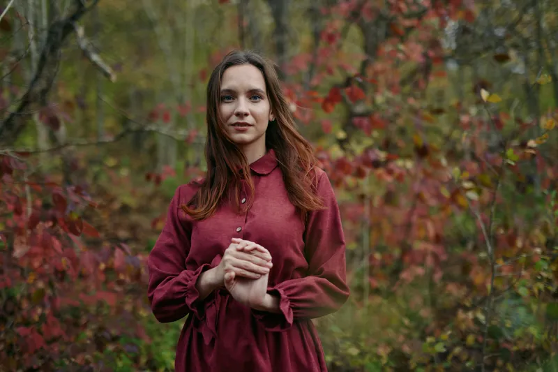 Young woman in a burgundy dress stands among autumn woodland foliage, hands gently clasped, natural expression and soft lighting highlighting a candid portrait that emphasizes authenticity and