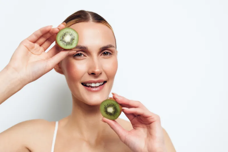 Smiling young woman holds kiwi slices like eye masks for skincare and beauty care on a clean light background Fresh fruit wellness concept for hydration spa routine and hydration
