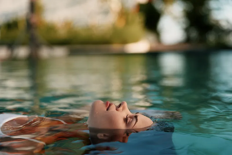 Relaxed woman floating on water surface with eyes closed, peaceful expression, soft natural light, outdoor pool setting, calm atmosphere, summer lifestyle moment