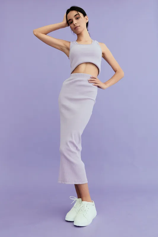 Slim woman wearing a lavender ribbed crop top and skirt set posing against a solid purple background, styled with white sneakers in modern casual fashion.