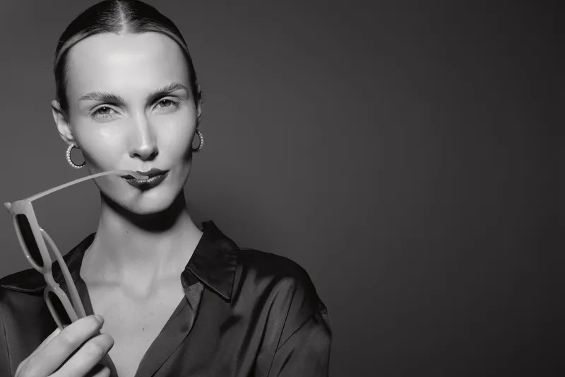 Slim Caucasian woman with pale skin and slicked back hair holds sunglasses near lips in black shirt on dark background. Concept of fashion elegance and minimalism in portrait photography.