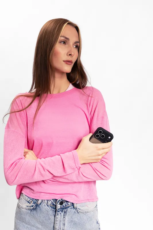 Young woman in pink sweater holding smartphone looks away thoughtfully. Casual style with long straight hair and denim jeans on white background. Concept of communication and contemplation.