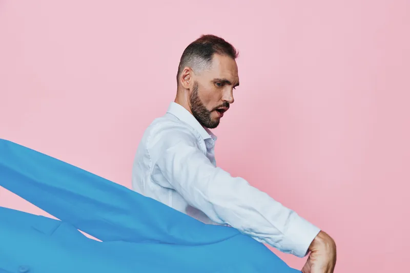 Professional man in business attire stands before a solid pink background, his face showing focus and determination as he grasps a vivid blue fabric, creating dynamic motion.