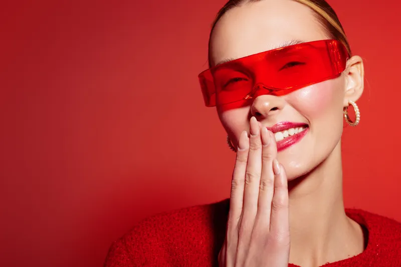 Smiling woman with red sunglasses and red sweater covers mouth with hand on red background. Concept of fashion, style, beauty, and joyful emotions in studio portrait photography.