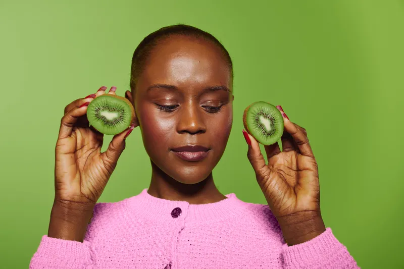 Dark skinned African woman in pink sweater holds two kiwi halves close to face on green background. Concept of healthy lifestyle, natural fruits, and wellness with fresh organic food.