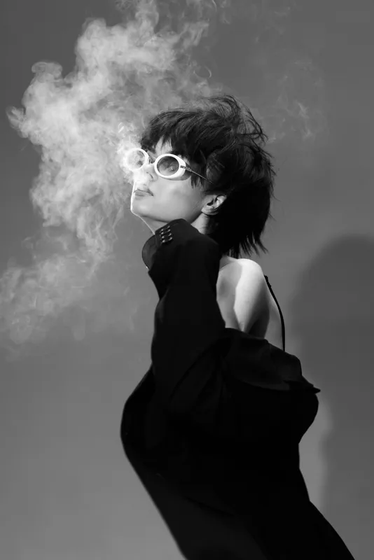 Black and white artistic portrait of a woman in sunglasses exhaling smoke with a stylish outfit.